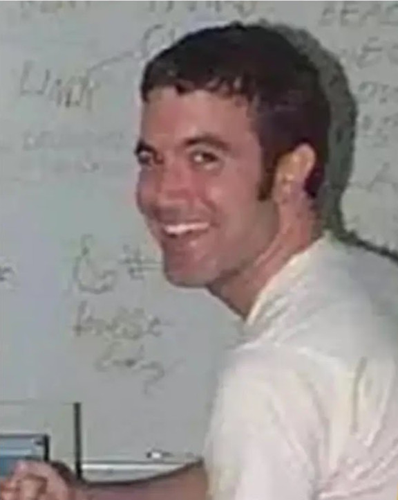 a white guy with reddish hair wearing a white shirt looks back over his shoulder